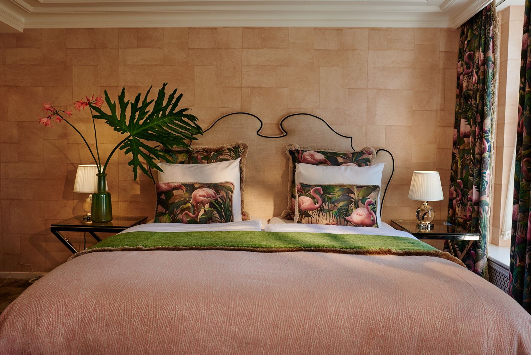 A hotel room with a bed, two nightstands, a green plant, and a tropical-themed curtain.