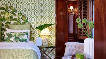Una habitación de hotel con papel tapiz verde y blanco con motivos, una cama con ropa de cama verde y blanca, un sillón blanco, una mesita de noche con una lámpara y una planta grande.