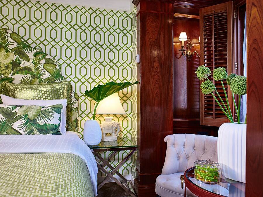 Una habitación de hotel con papel tapiz verde y blanco con motivos, una cama con ropa de cama verde y blanca, un sillón blanco, una mesita de noche con una lámpara y una planta grande.