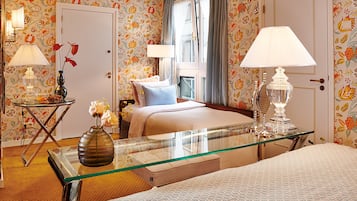 Habitació d'hotel amb paper pintat floral, un llit amb almoades, una taula de vidre amb un ram i dues làmpades.