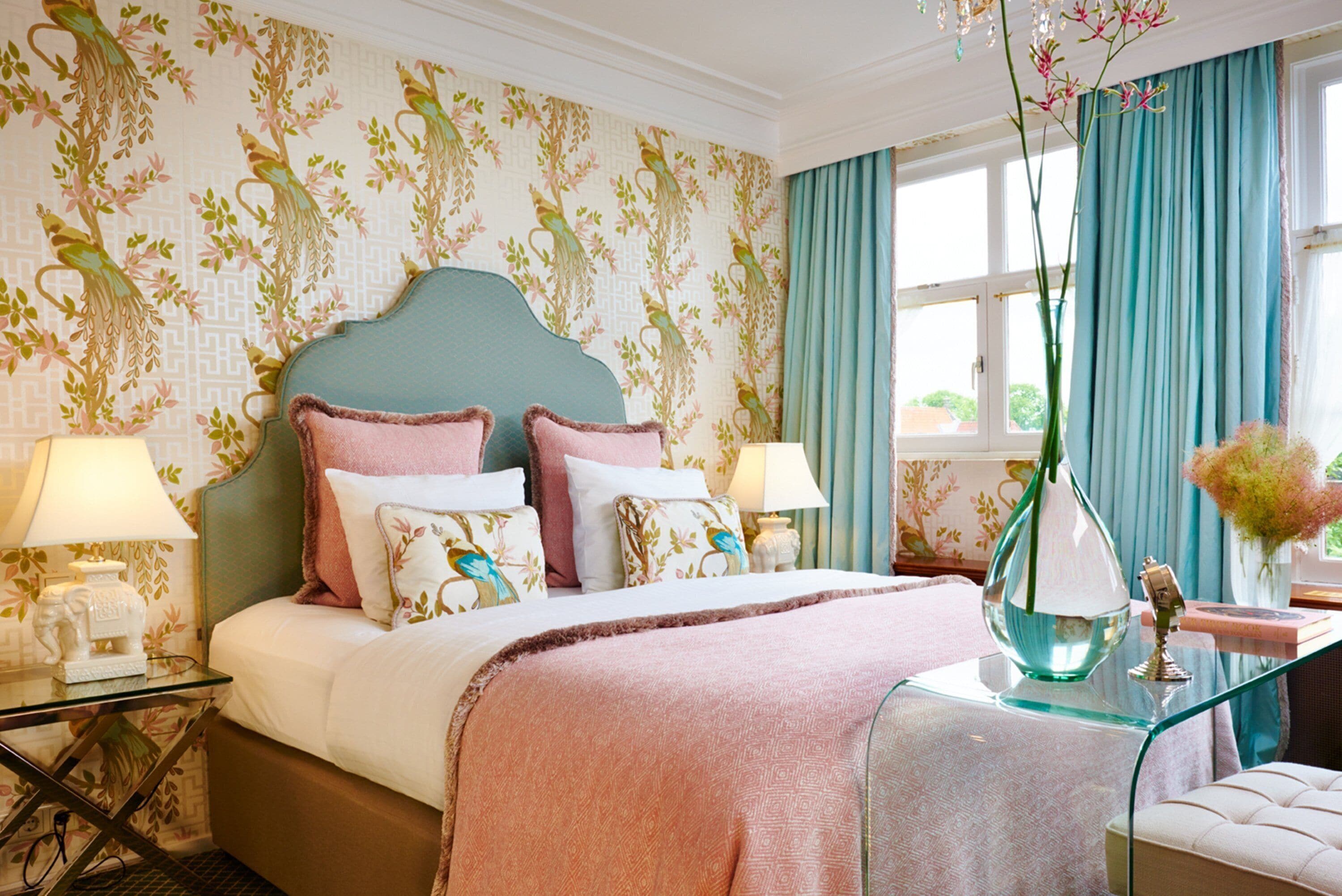A hotel room with a bed, bedside tables, lamps, a vase with flowers, and a window with curtains.