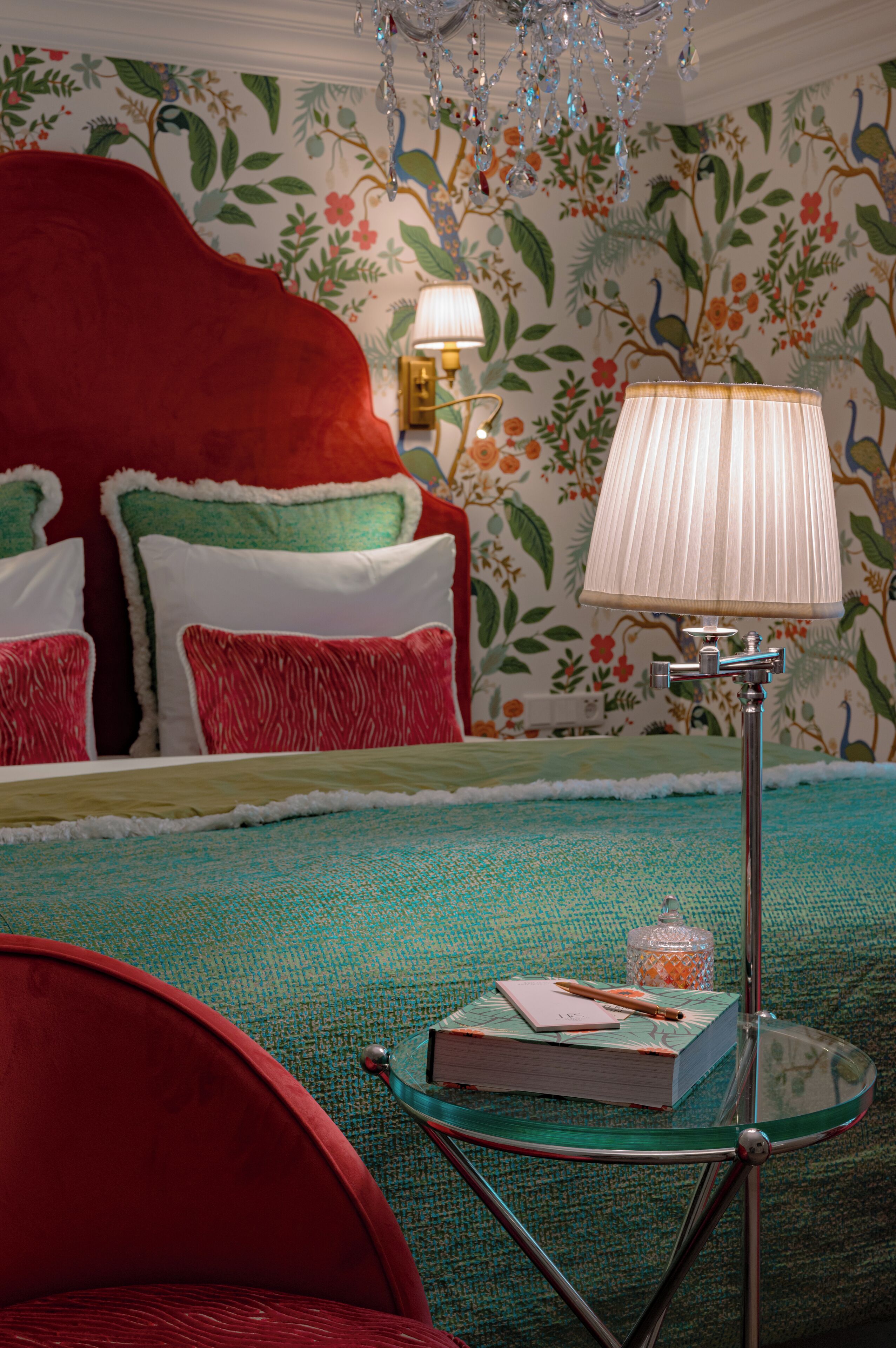 A bedroom with a red upholstered headboard, floral wallpaper, a glass table with a book and a lamp, and a teal bedspread.