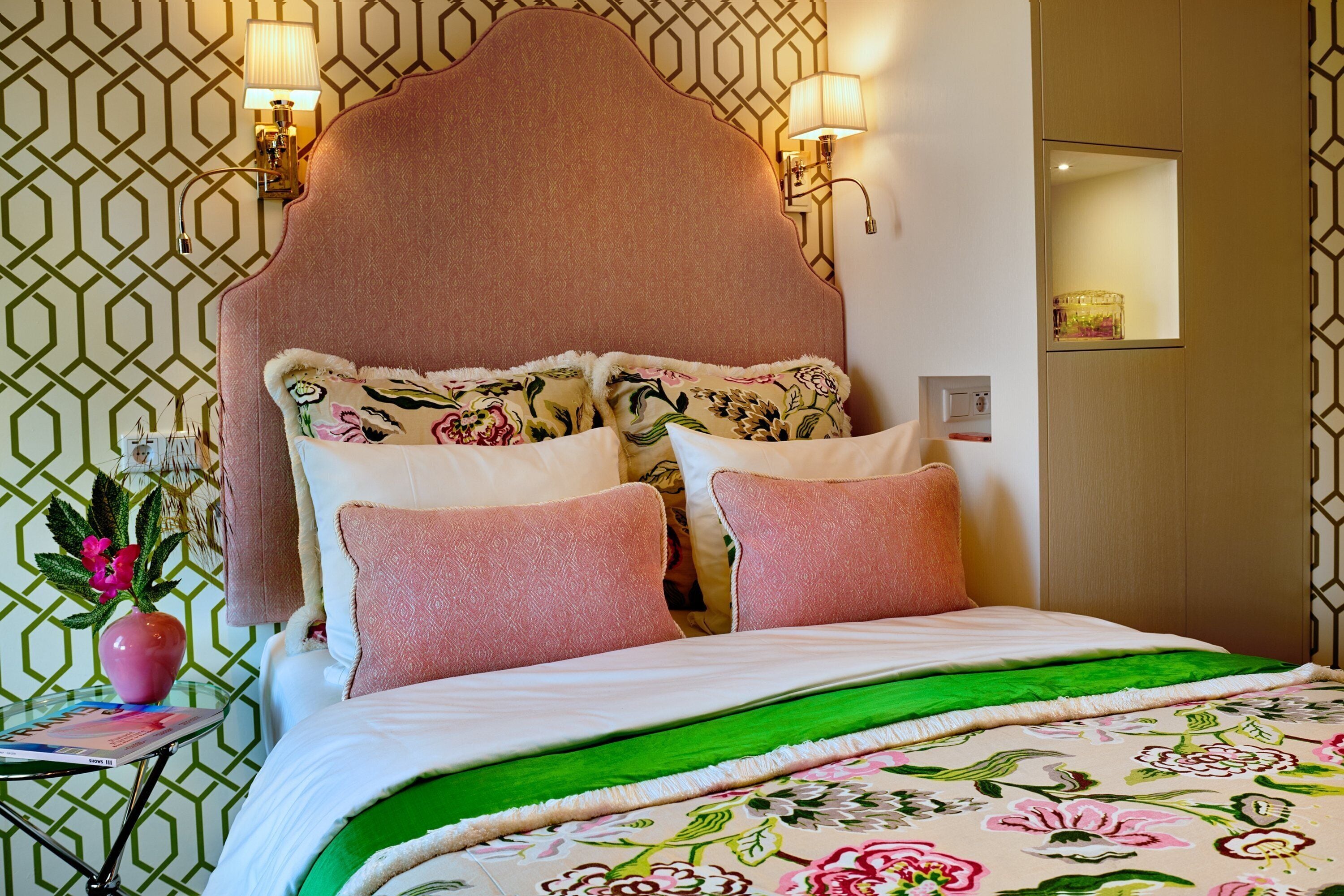 Una habitación de hotel con una cama grande, ropa de cama floral, un plaid verde y un papel tapiz con motivos geométricos.