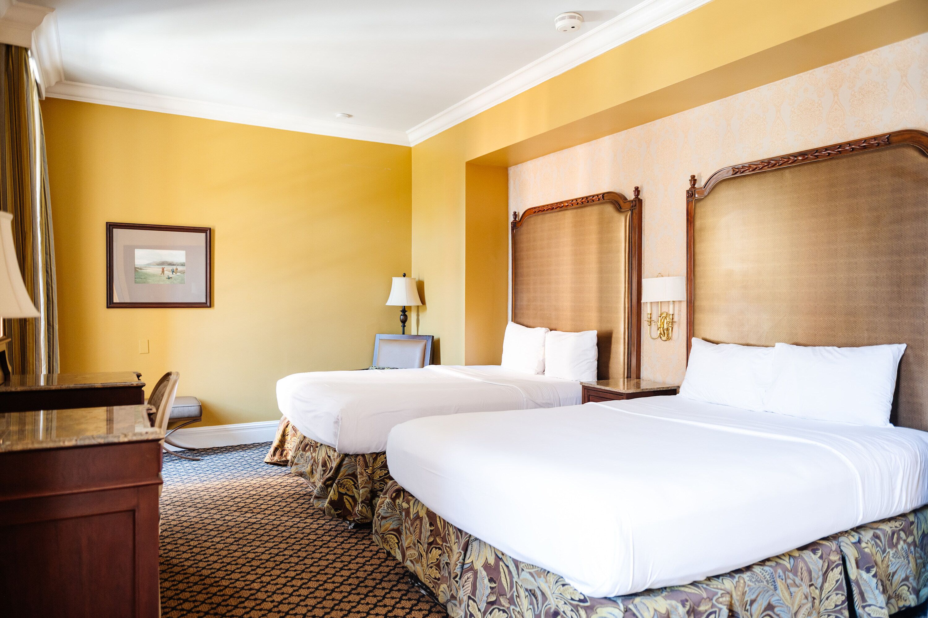 A hotel room with two beds, a desk, a chair, a lamp, and a framed picture on the wall.