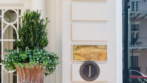 Fachada de un edificio histórico con una placa que indica que es un hotel recomendado, una maceta grande con plantas y una entrada empaveda de ladrillo.