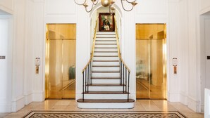 Una imponente escalera con candelabro, flanqueada por dos ascensores con puertas doradas.