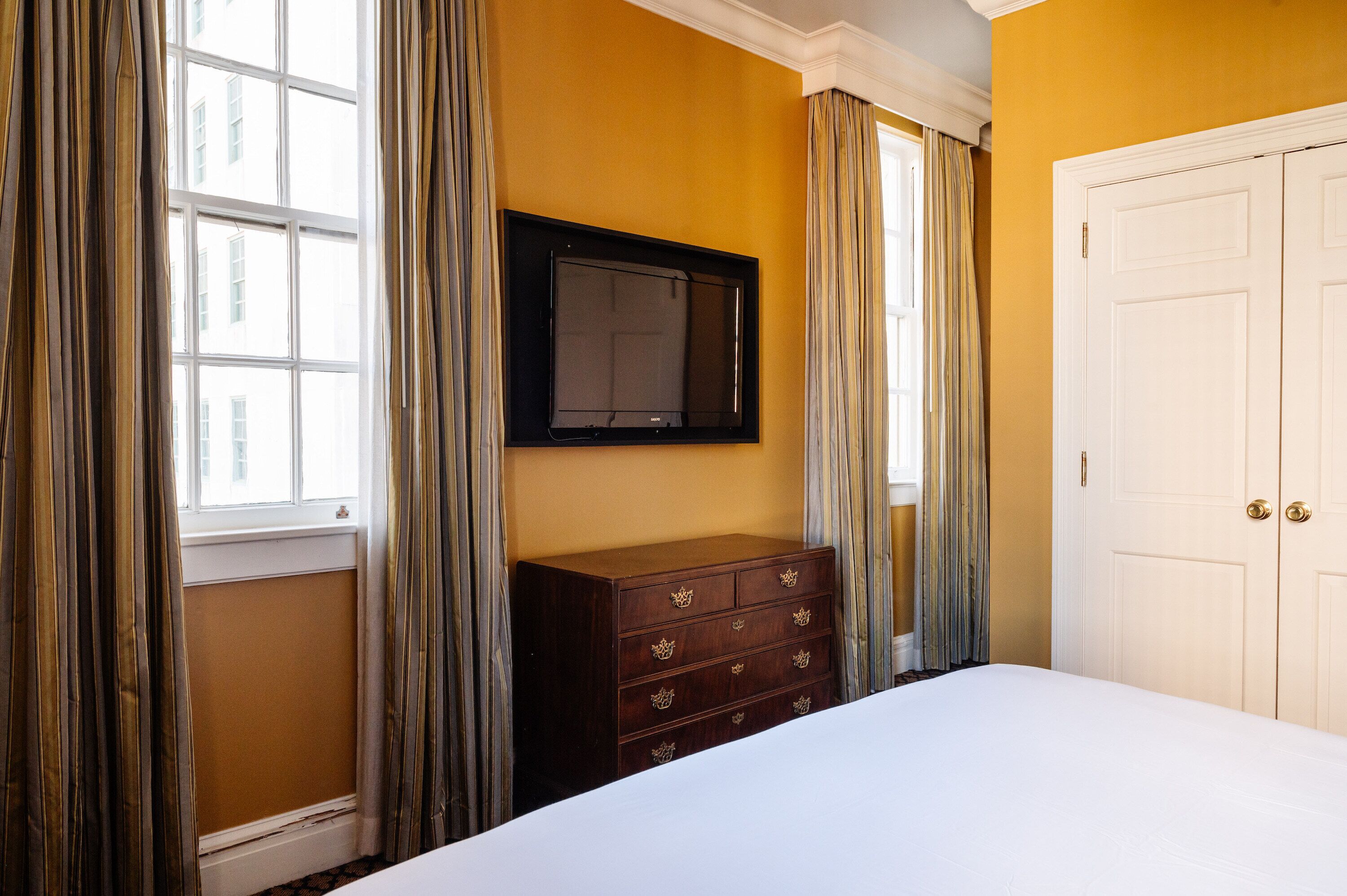 A hotel room with a large bed, a dresser with crown handles, a flat-screen TV mounted on the wall, and a door with a gold handle.