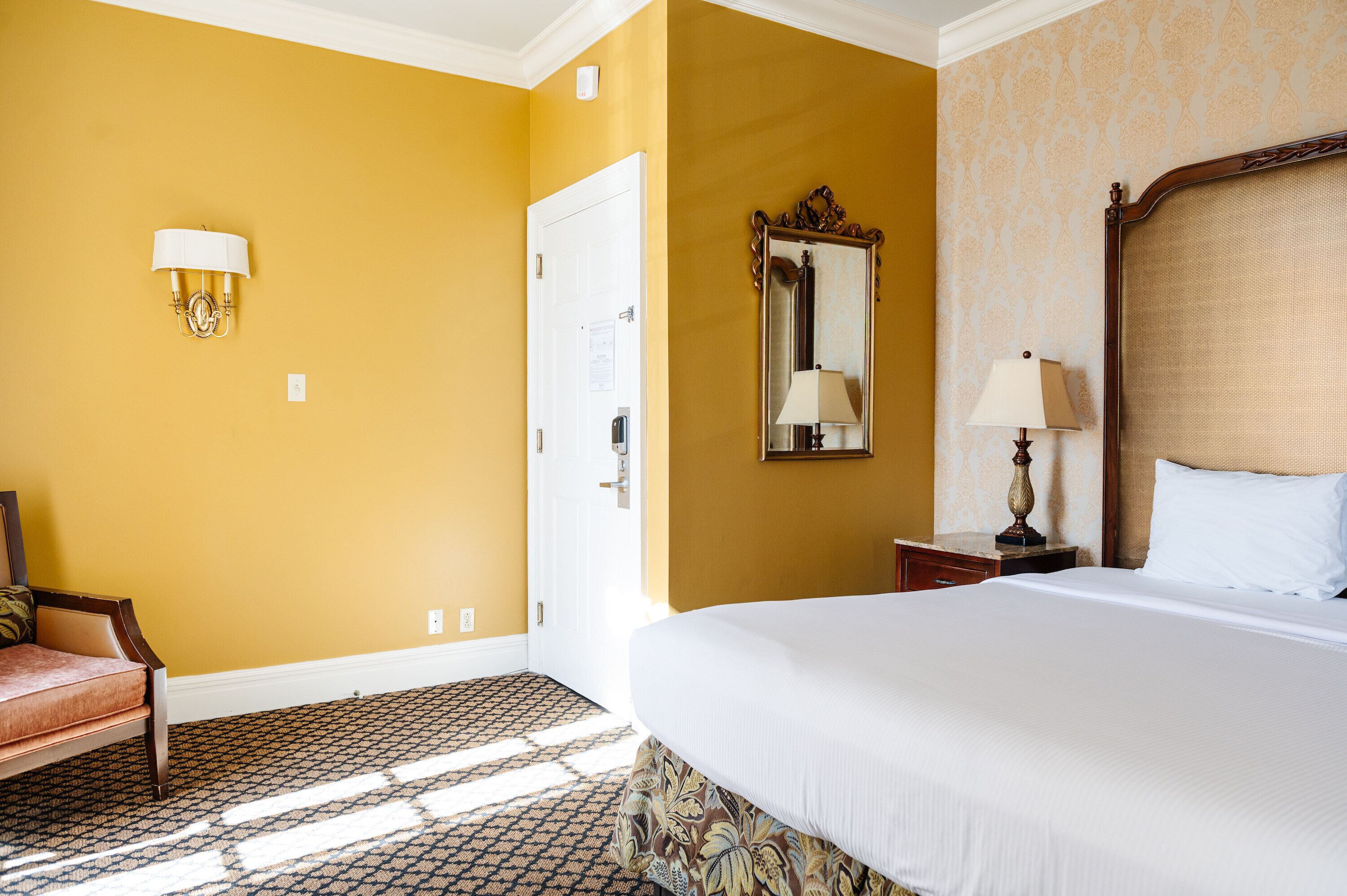 Une chambre d’hôtel avec un grand lit, une chaise, une lampe fixée au mur, un miroir et une porte.