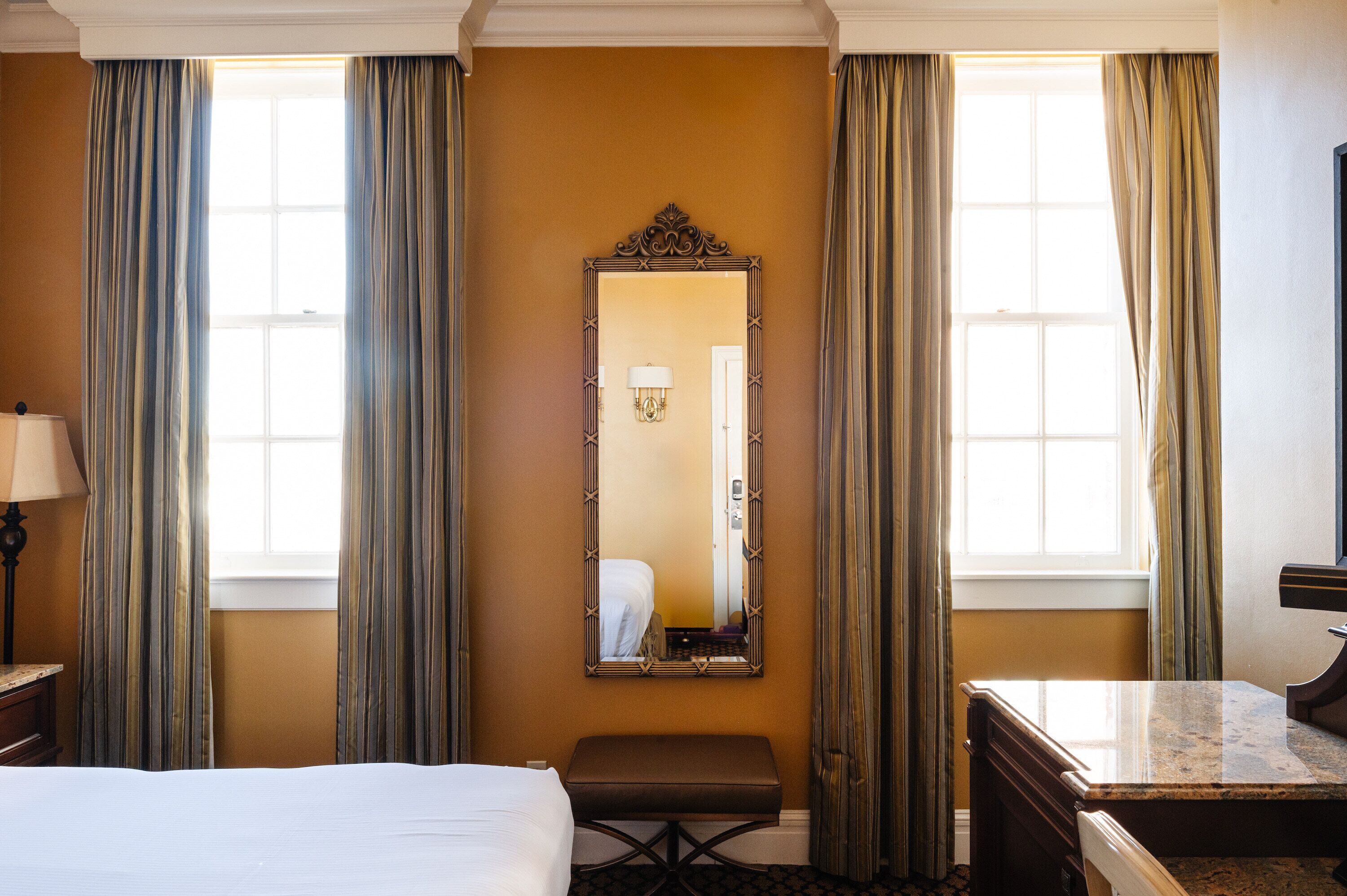 A hotel room with a bed, a bench, a desk, a mirror, and curtains.