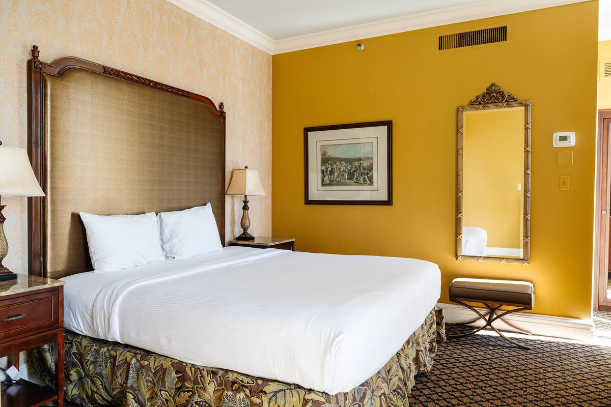 A hotel room with a large bed, a bedside table, a lamp, a framed picture on the wall, and a mirror.