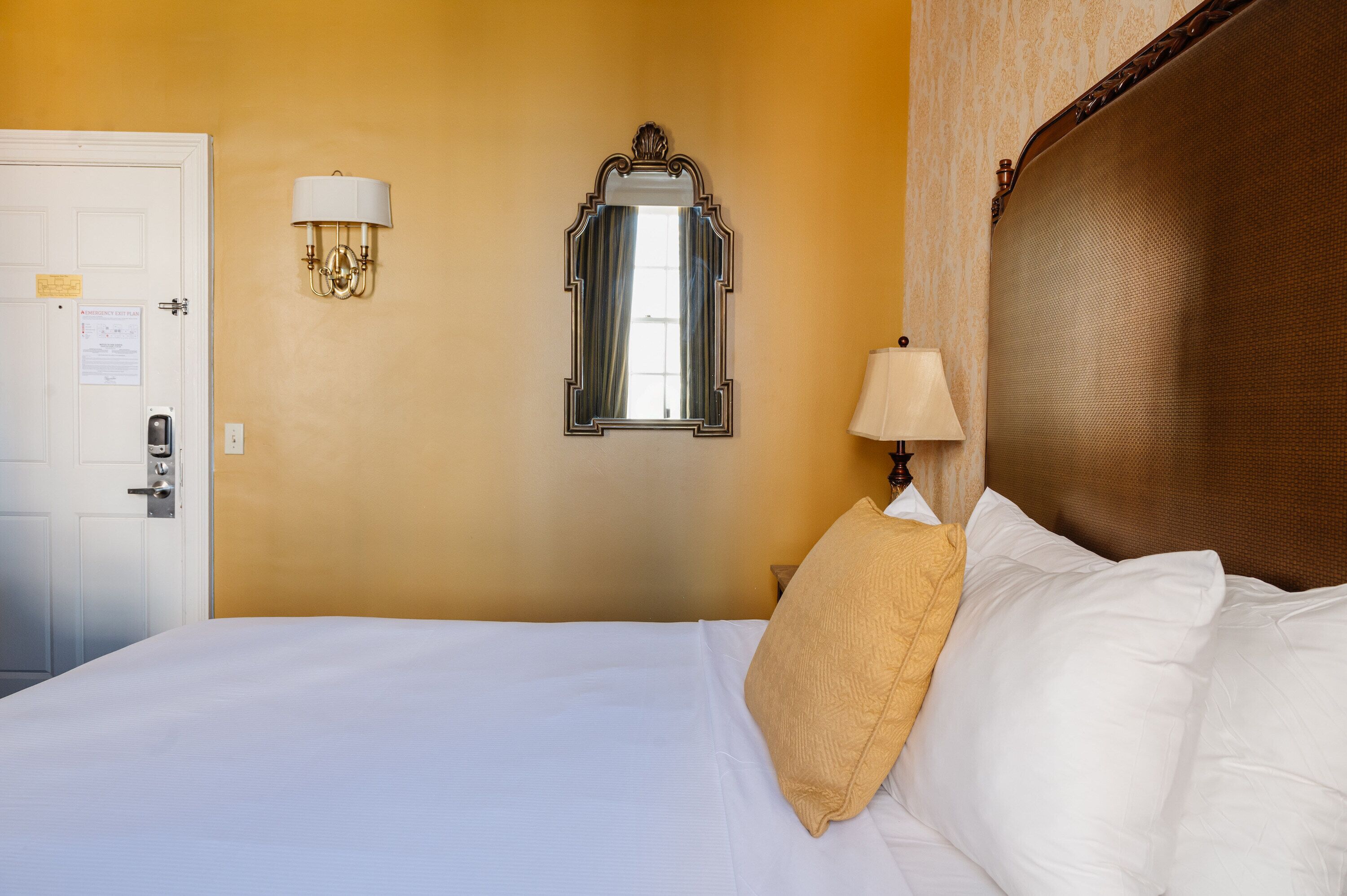 Una habitación de hotel con cama, una puerta, un espejo montado en la pared y una lámpara de pared.