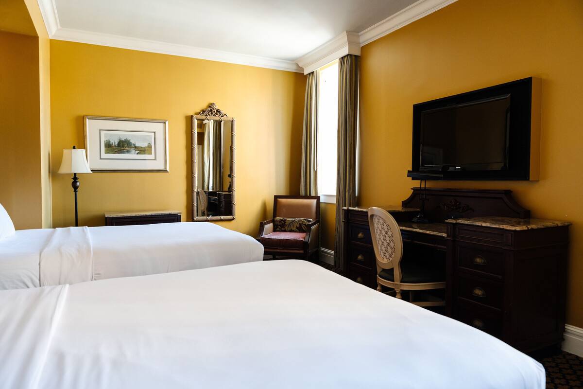 A hotel room with two beds, a desk, a chair, a dresser, a television, and a window with curtains.