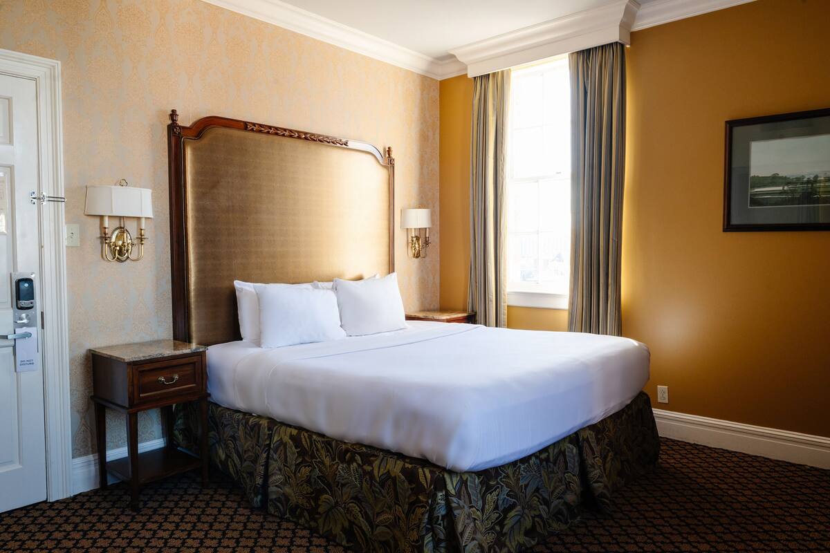 A hotel room with a large bed, two bedside tables with lamps, a door with a lock, and a window with curtains.
