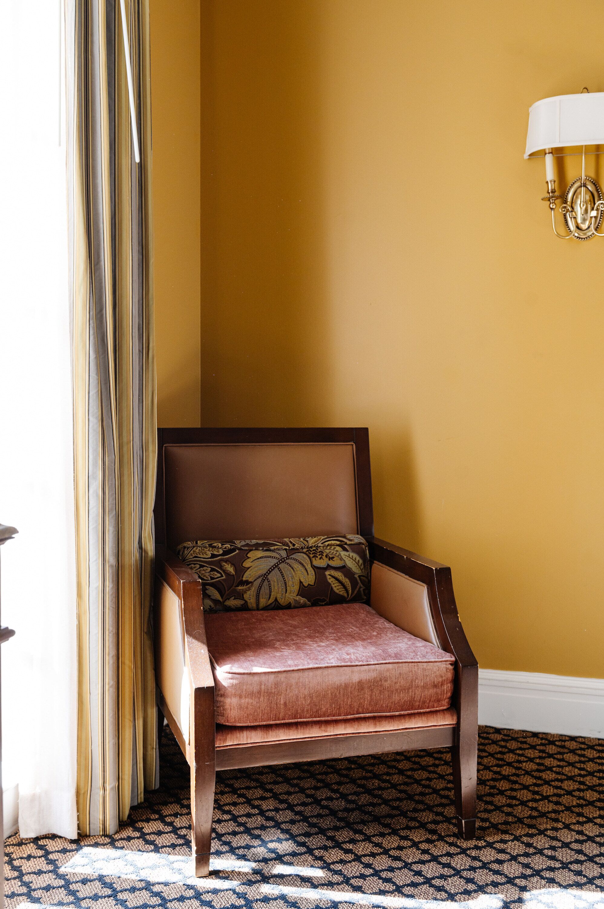 Una habitación de esquina con alfombra estampada, un sillón tapizado en marrón y un cojín floral, además de una lámpara fijada a la pared.