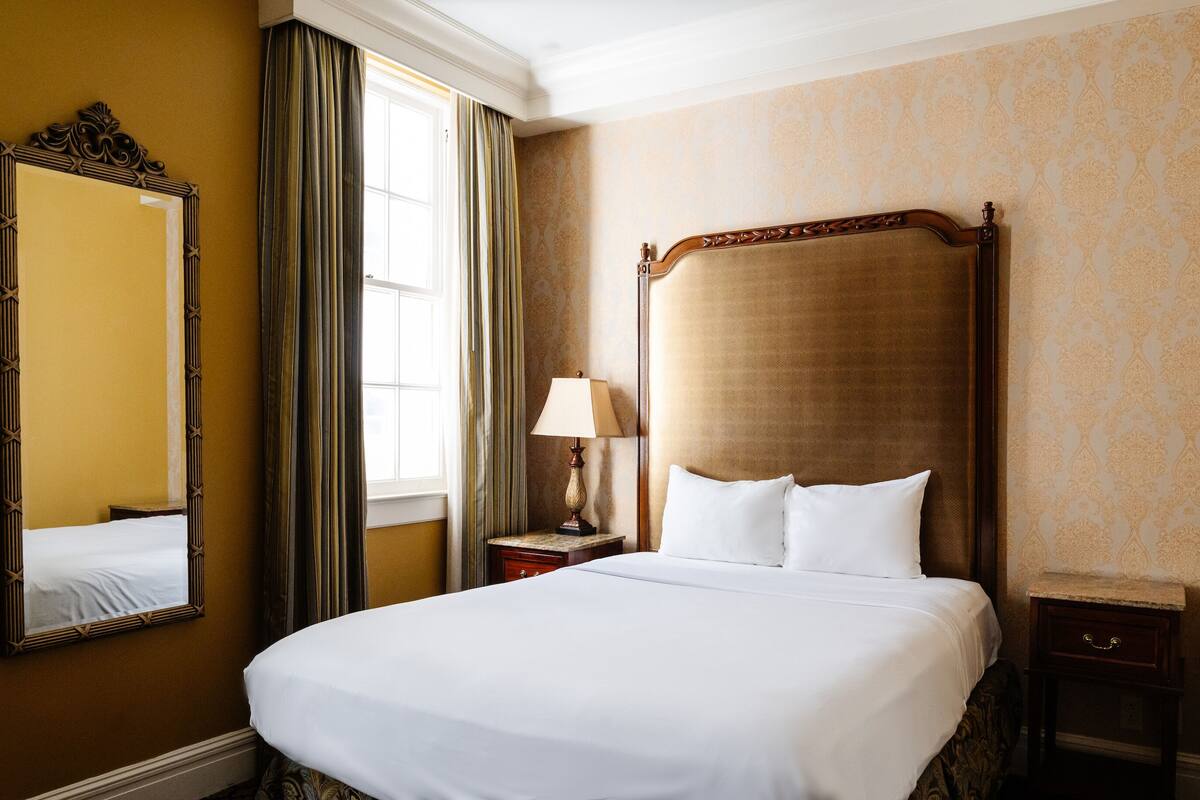A hotel room with a large bed, a nightstand with a lamp, a mirror, and a window with curtains.