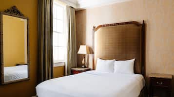 A hotel room with a large bed, a nightstand with a lamp, a mirror, and a window with curtains.