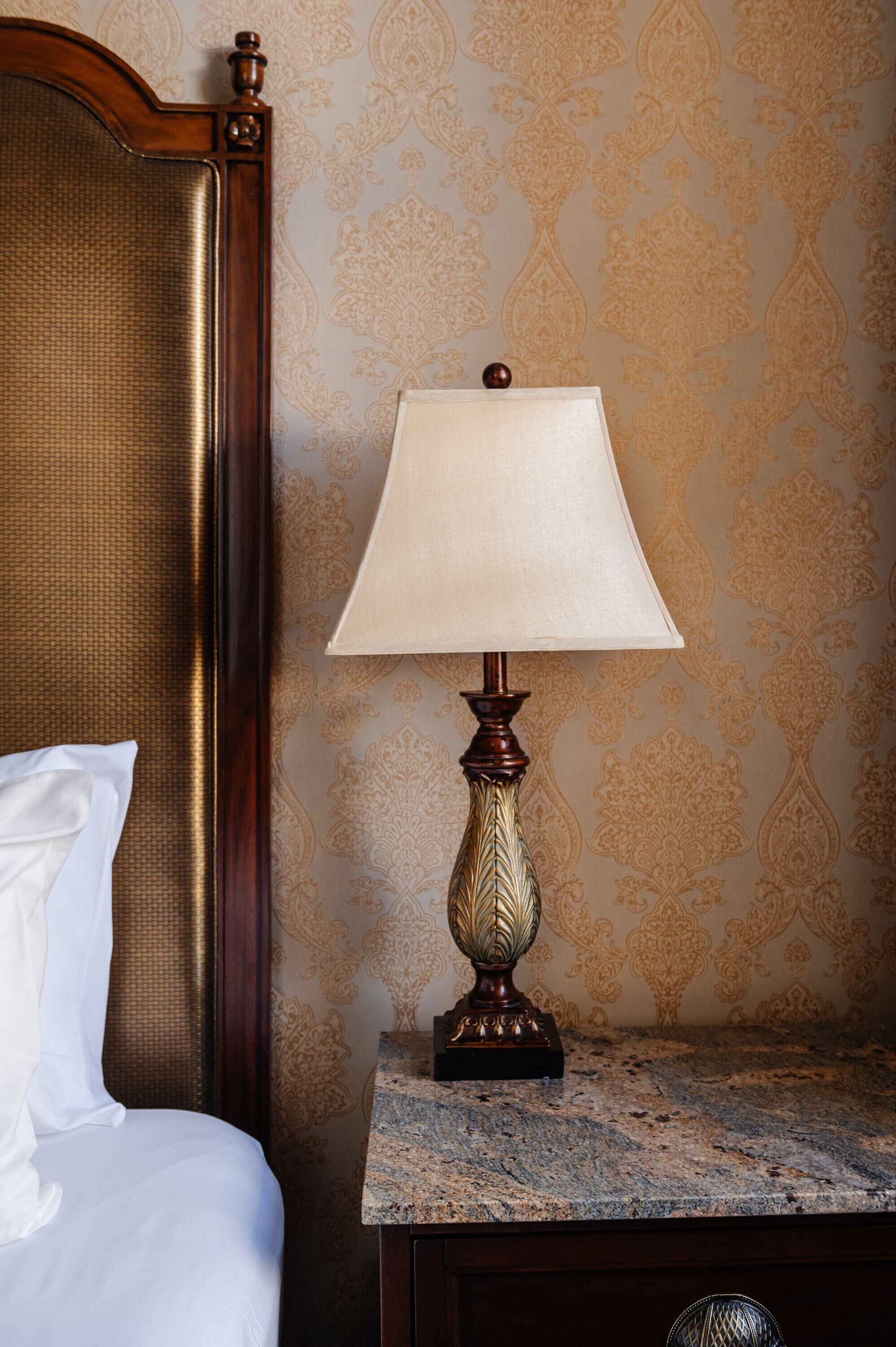 A bed with white linens, a wooden headboard, a marble nightstand, and a table lamp with a beige shade.