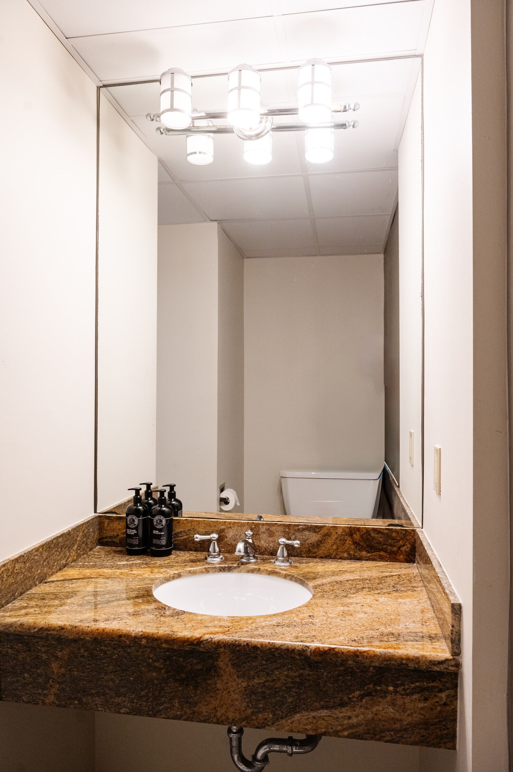 A bathroom with a granite countertop sink, a mirror, and a toilet visible in the reflection.