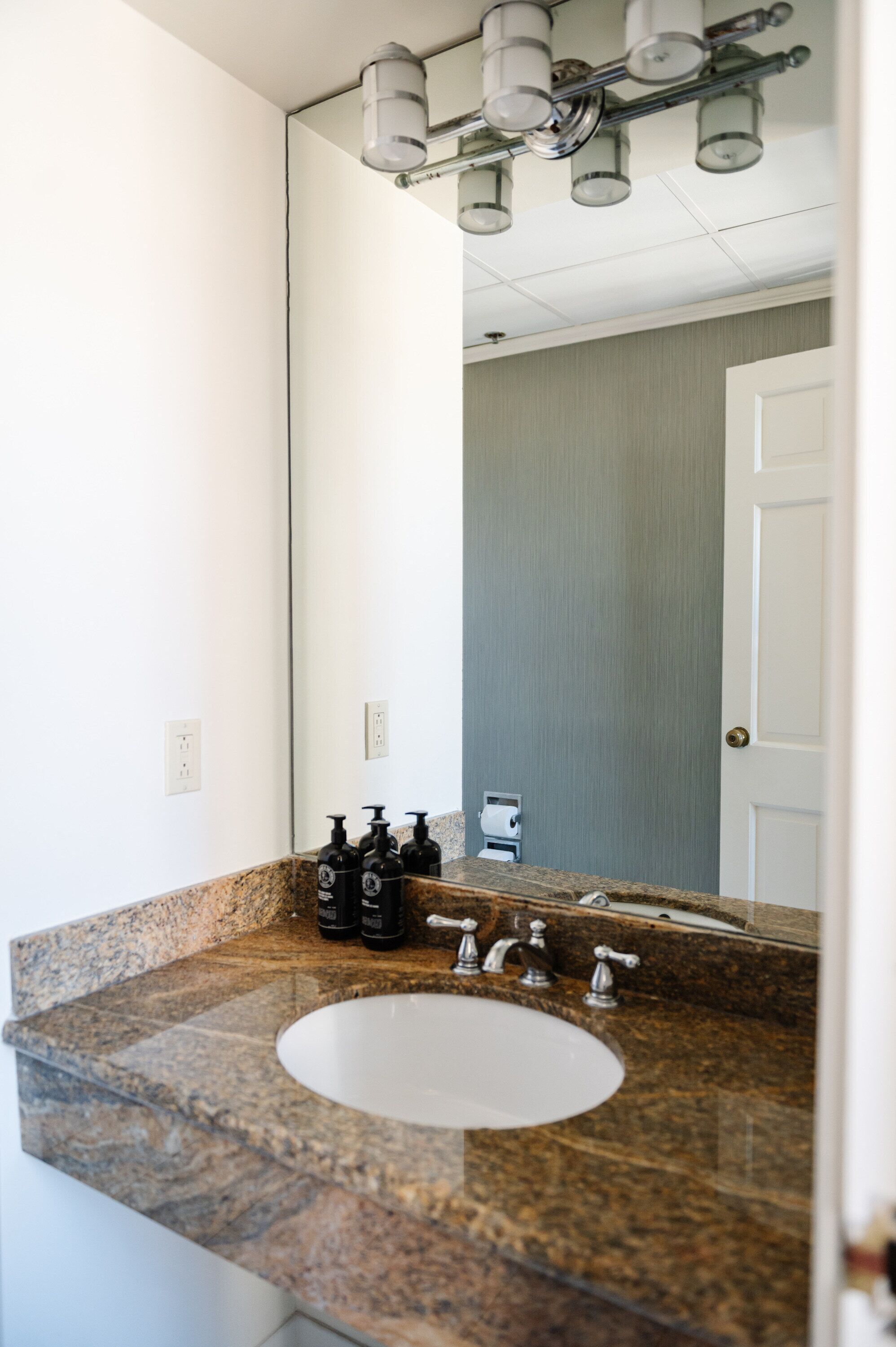 A bathroom with a granite countertop, a white sink, and a mirror. There are two black bottles on the counter, and a towel dispenser is visible in the mirror's reflection.