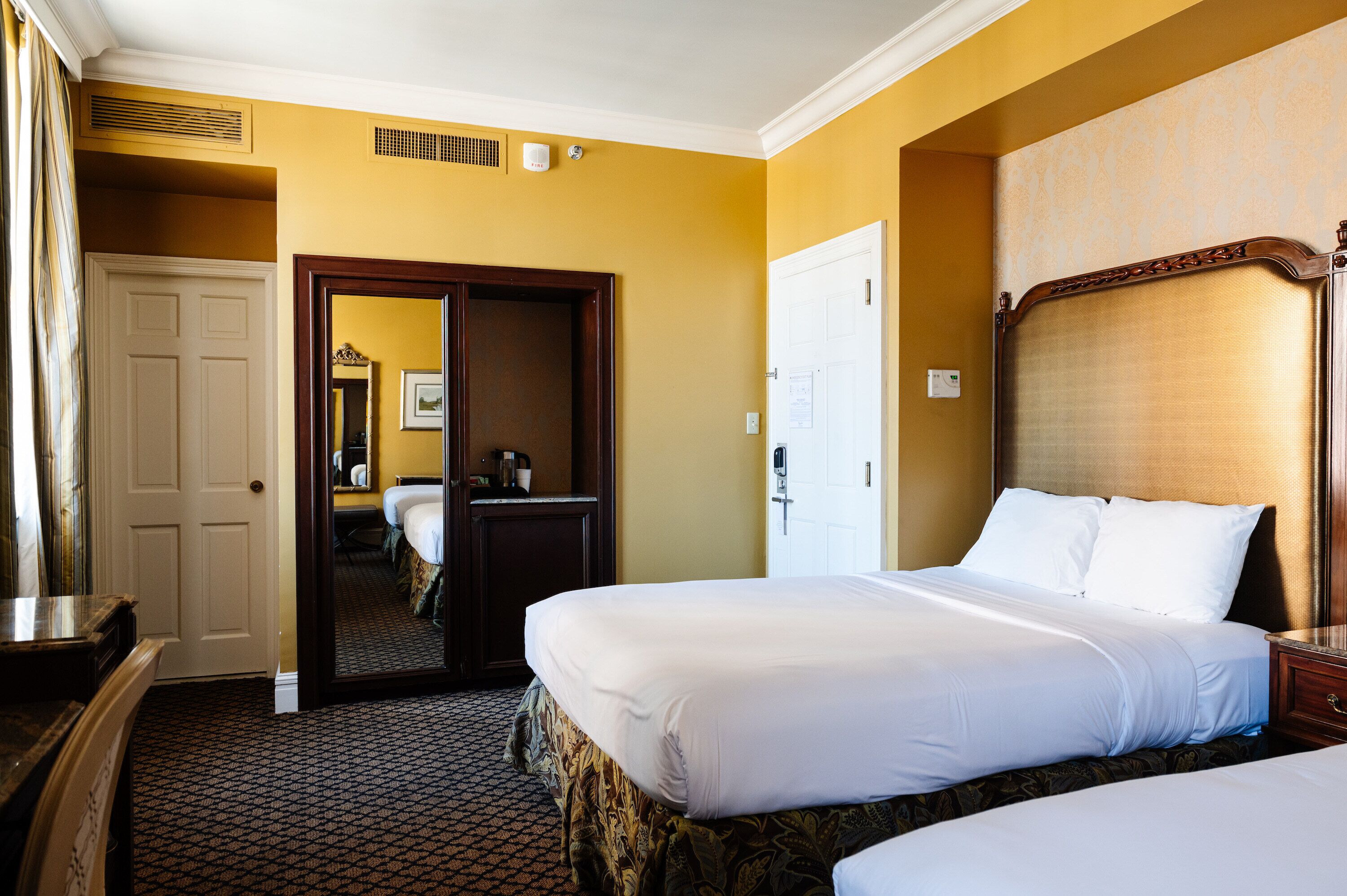 Une chambre d’hôtel avec deux lits, une tête de lit en bois, un miroir et une porte donnant sur une autre pièce.
