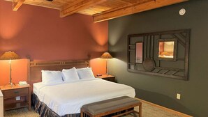 A bedroom with a wooden headboard, a large bed with white linens, two bedside lamps, a bench, and a wall-mounted mirror. - Crossroads Hotel and Event Center (Fort Dodge)