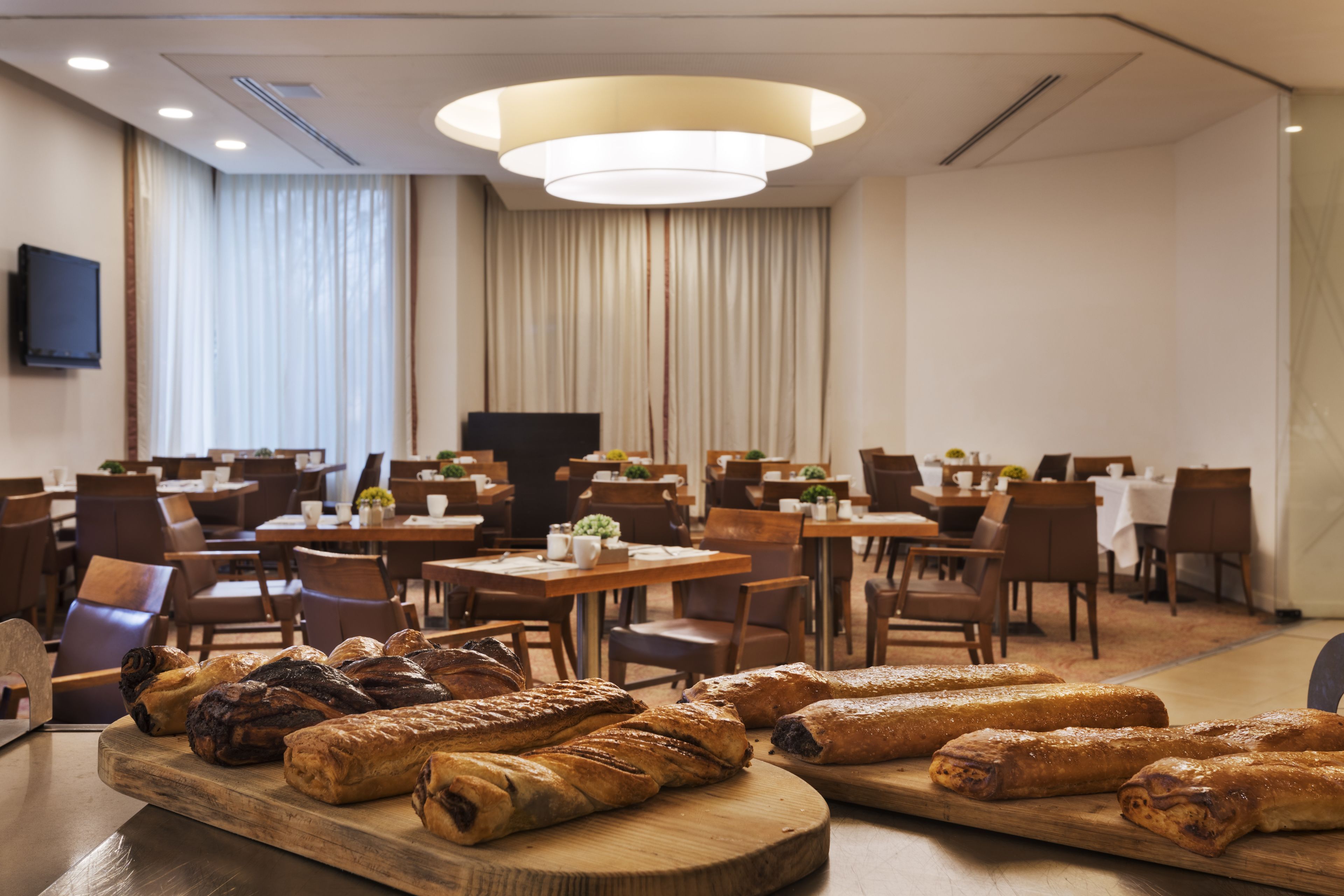 Un comedor de hotel con varias mesas y sillas, una amplia tabla de madera con pasteles, y un televisor montado en la pared.