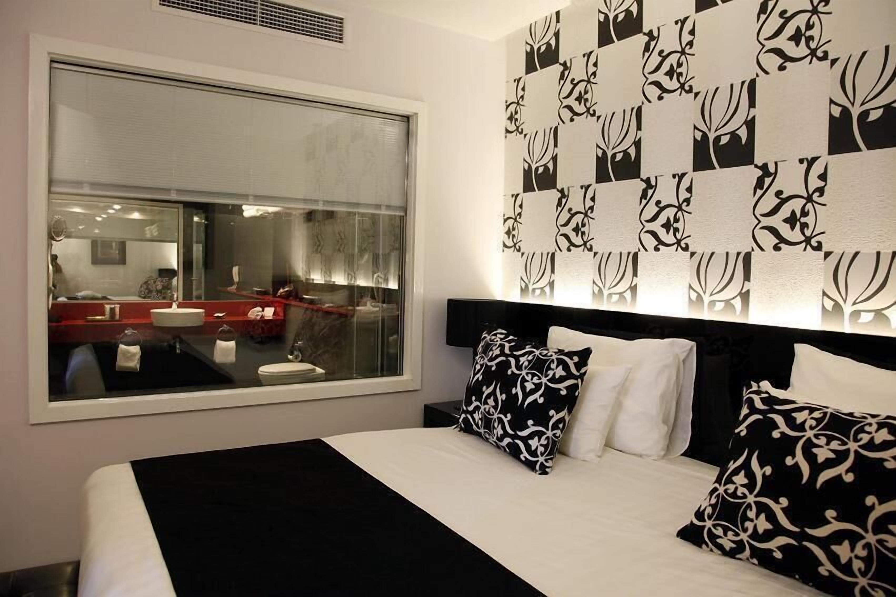 Habitació d'hotel moderna amb una finestra gran, un llit amb almoinos de disseny blanc i negre, i un mirall fixat a la paret.