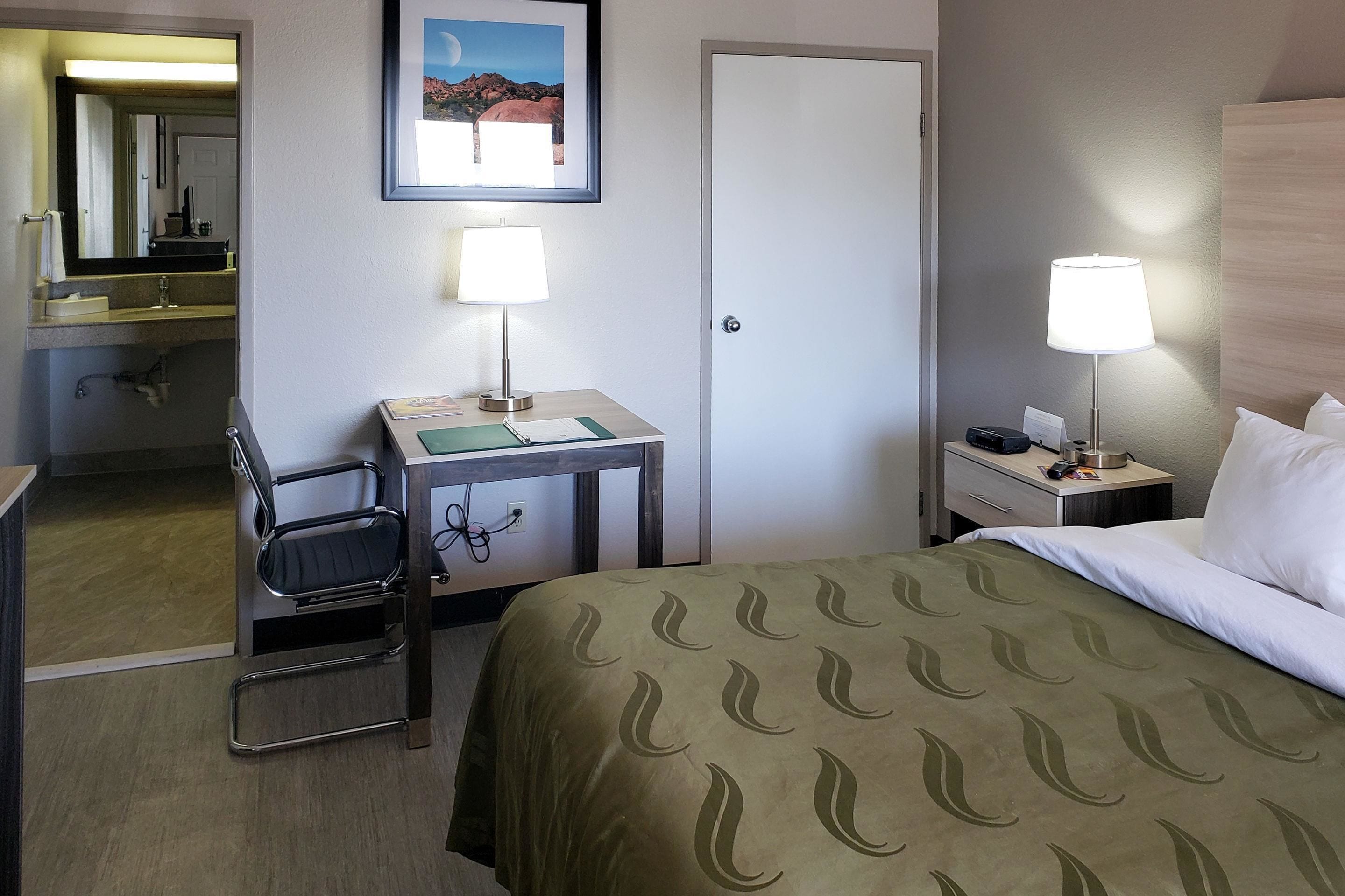 Habitación de hotel con cama, dos mesitas de noche con lámparas, un escritorio con silla, un espejo y una puerta.