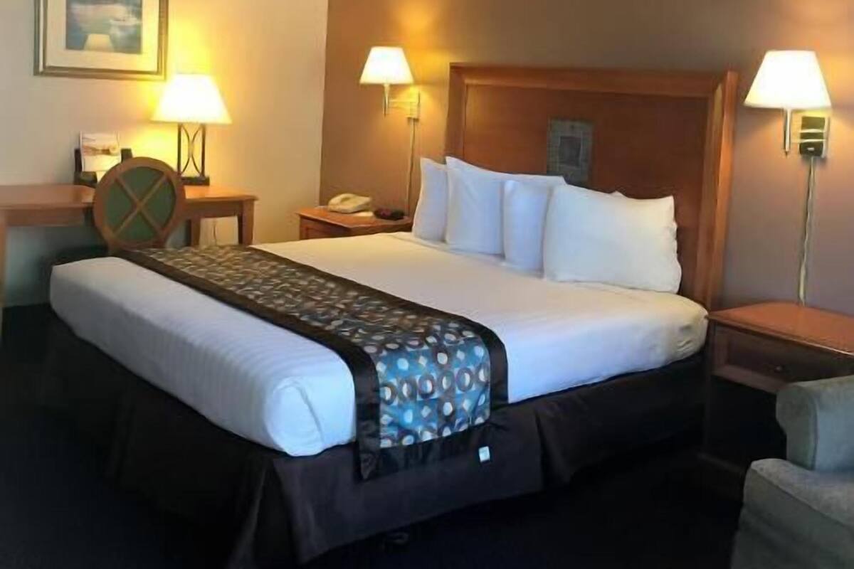 A hotel room with a large bed, two bedside lamps, a wooden headboard, a nightstand with a phone, and a framed picture on the wall.