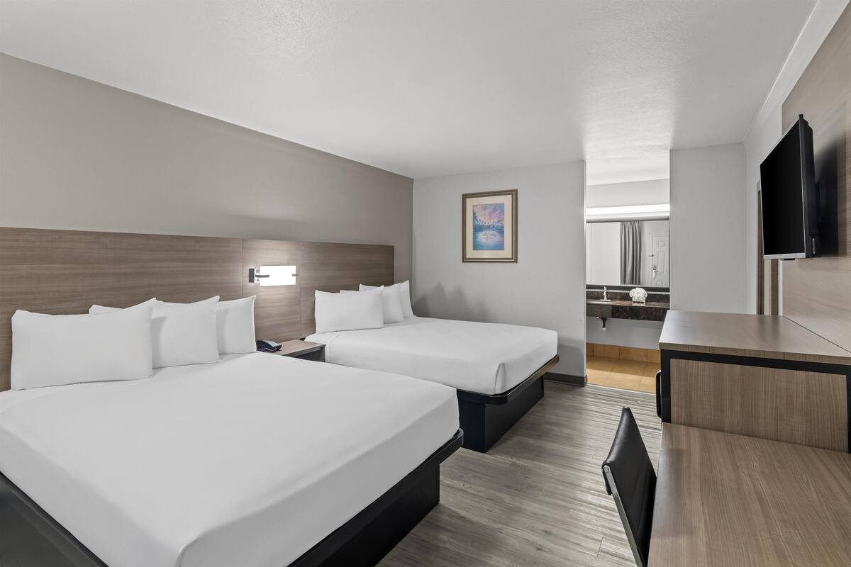 A hotel room with two beds, a desk, a TV, and a bathroom visible in the background.