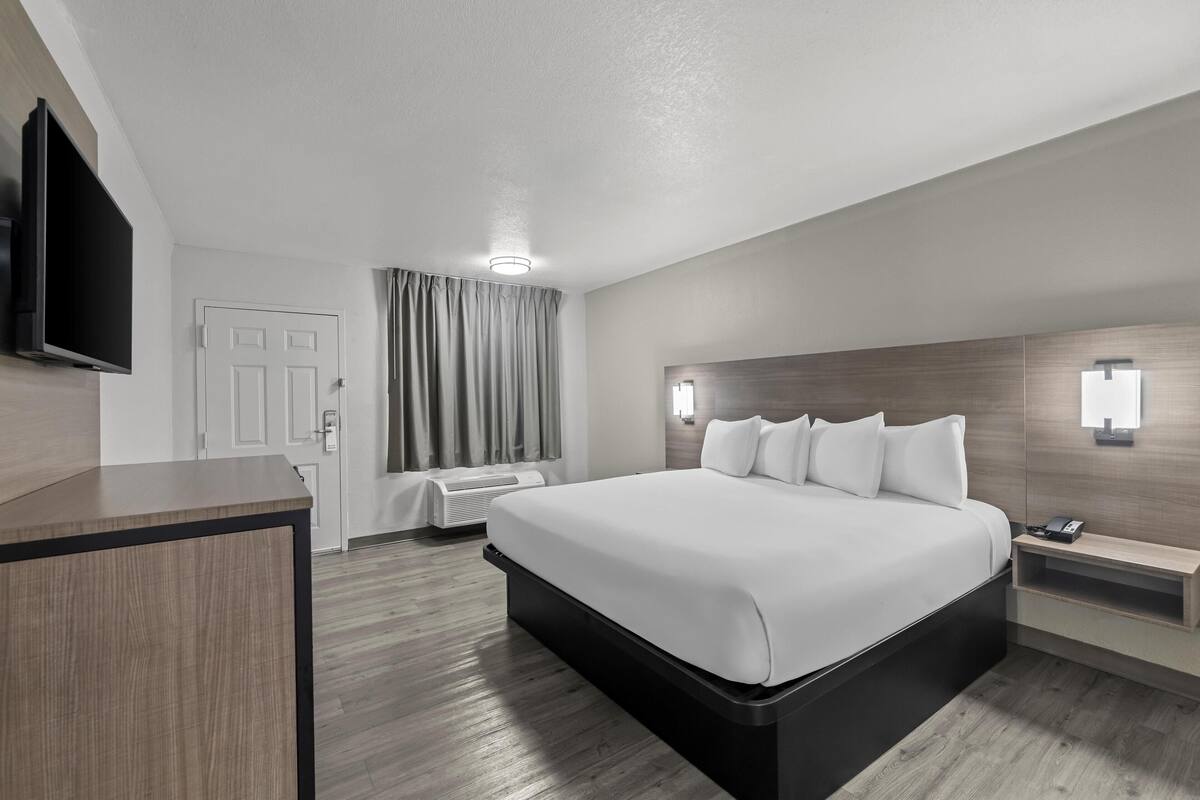 A modern hotel room with a large bed, a flat-screen TV mounted on the wall, a desk, and a bedside table with a lamp.