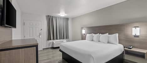 A modern hotel room with a large bed, a flat-screen TV mounted on the wall, a desk, and a bedside table with a lamp.