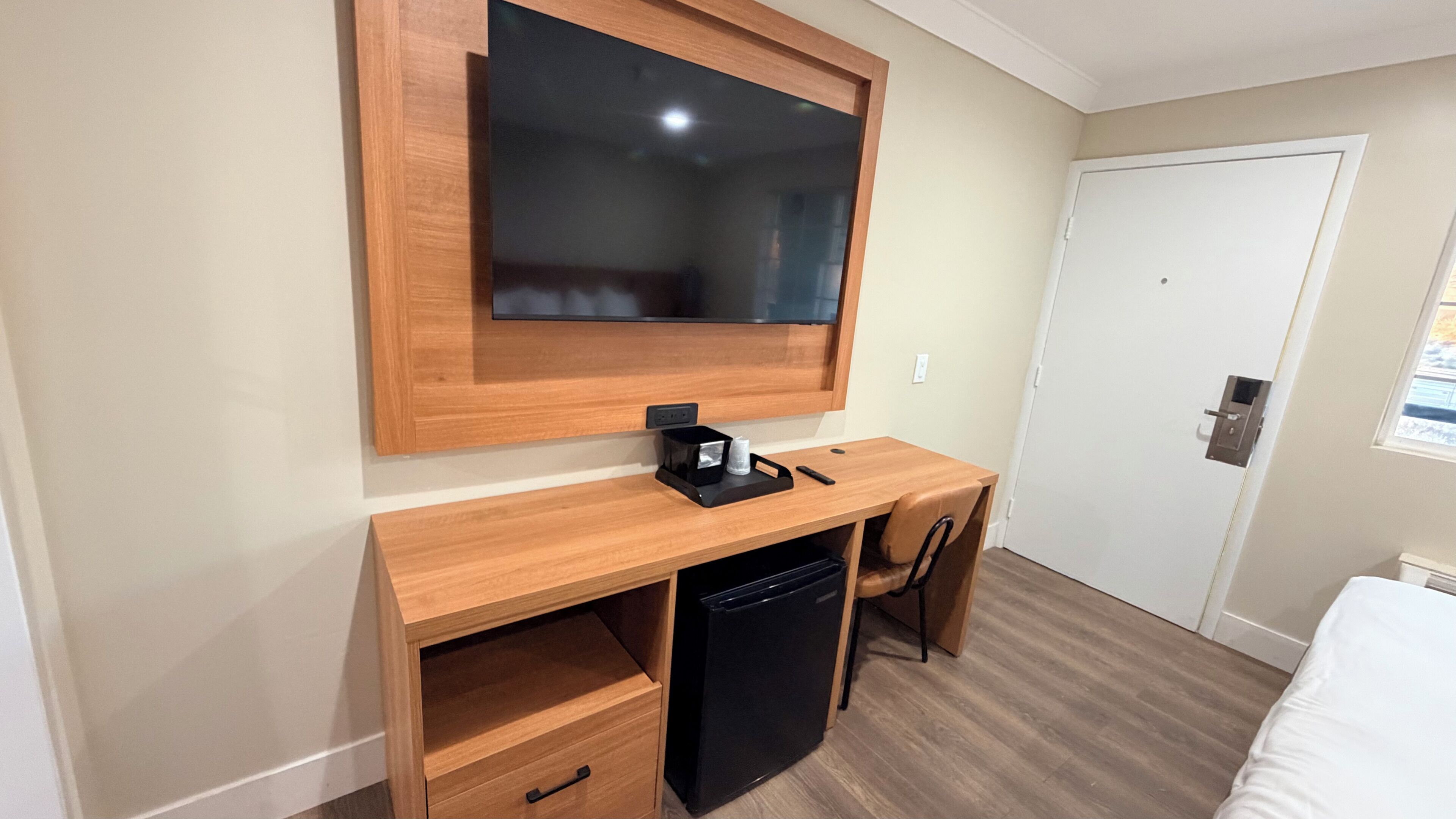 A hotel room with a wooden desk, a black refrigerator, a television mounted on the wall, and a white door.