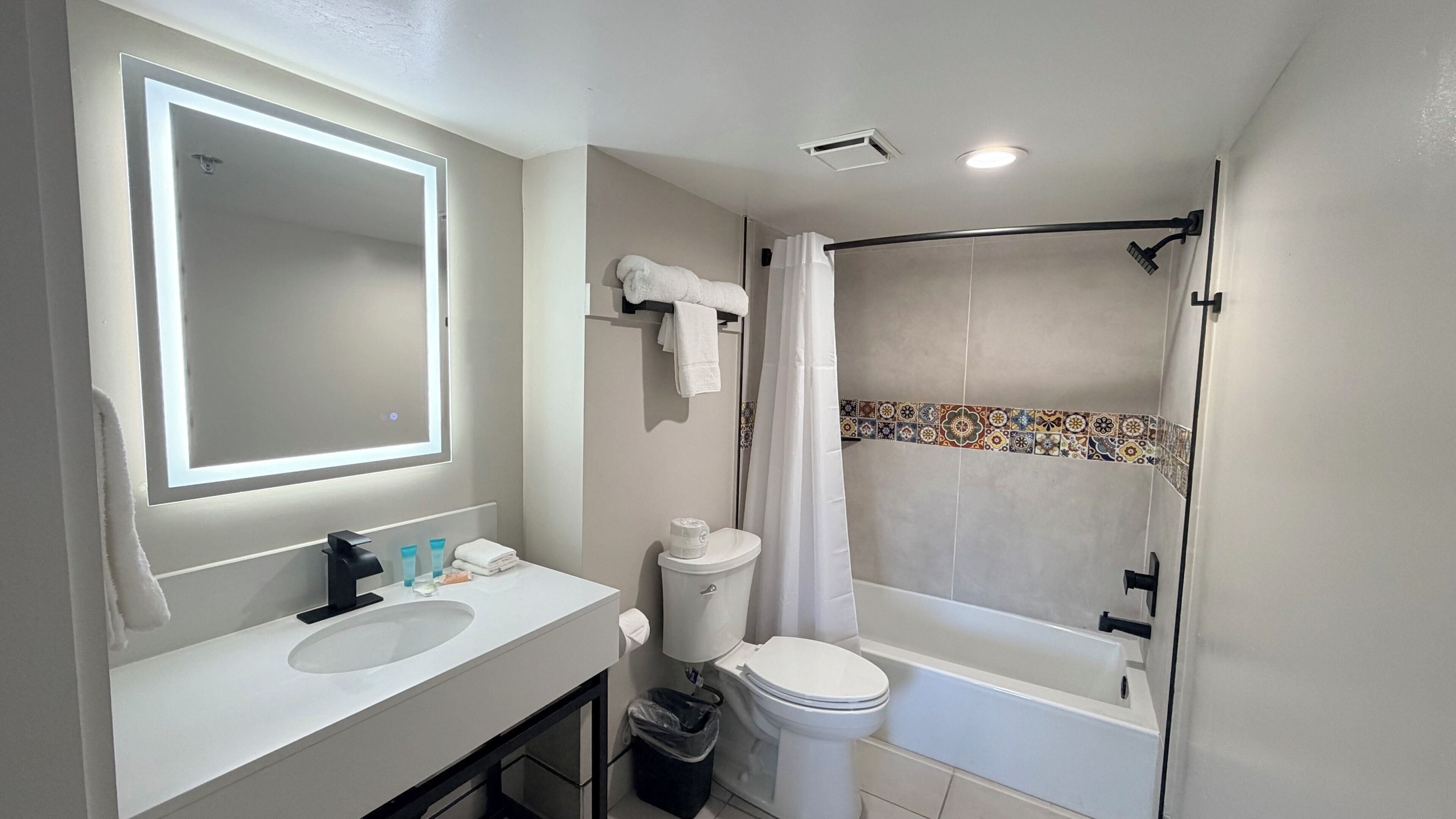 A bathroom with a sink, toilet, shower, and a mirror with lighting.