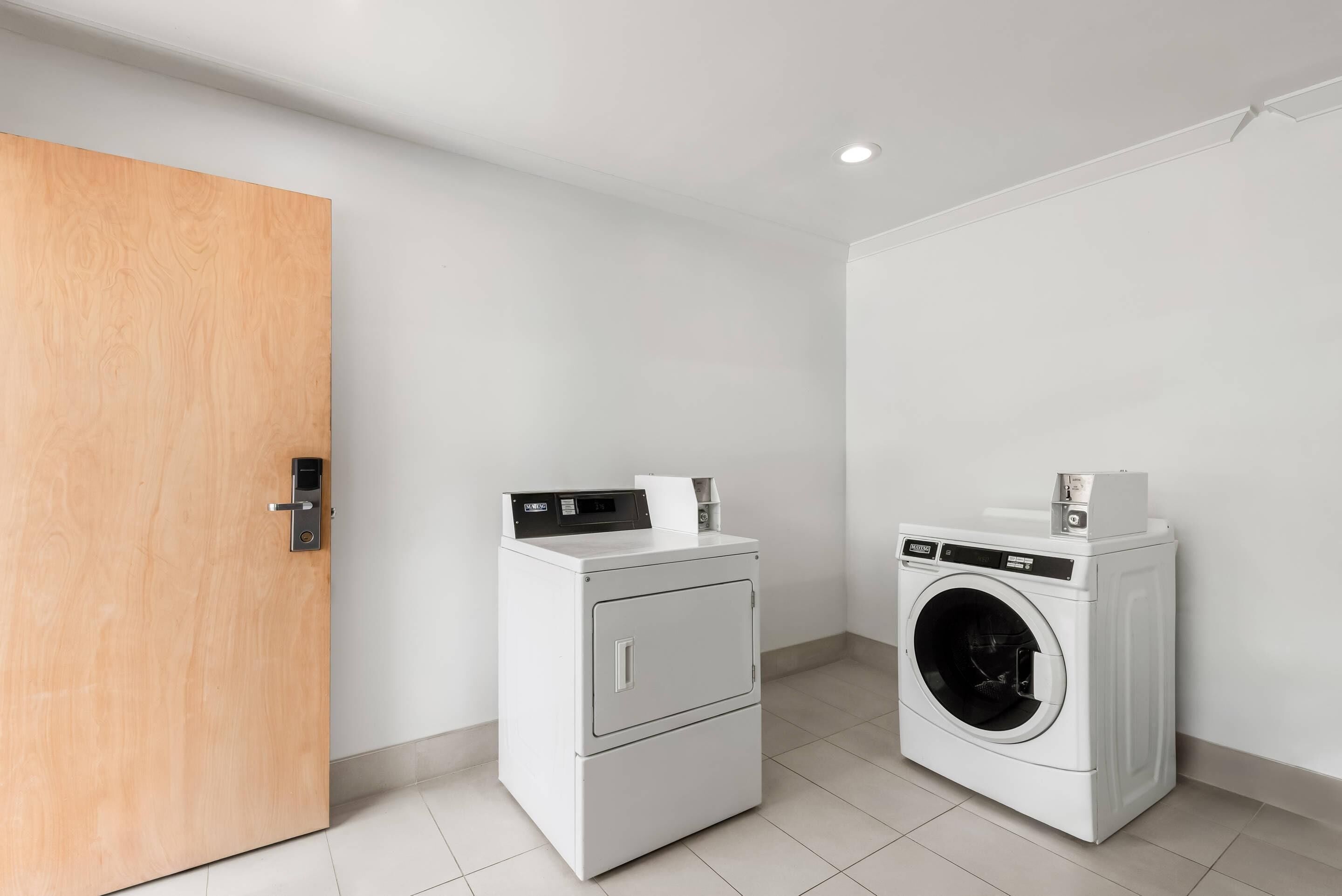 A laundry room with two white washing machines, a wooden door, and white tiled flooring.
