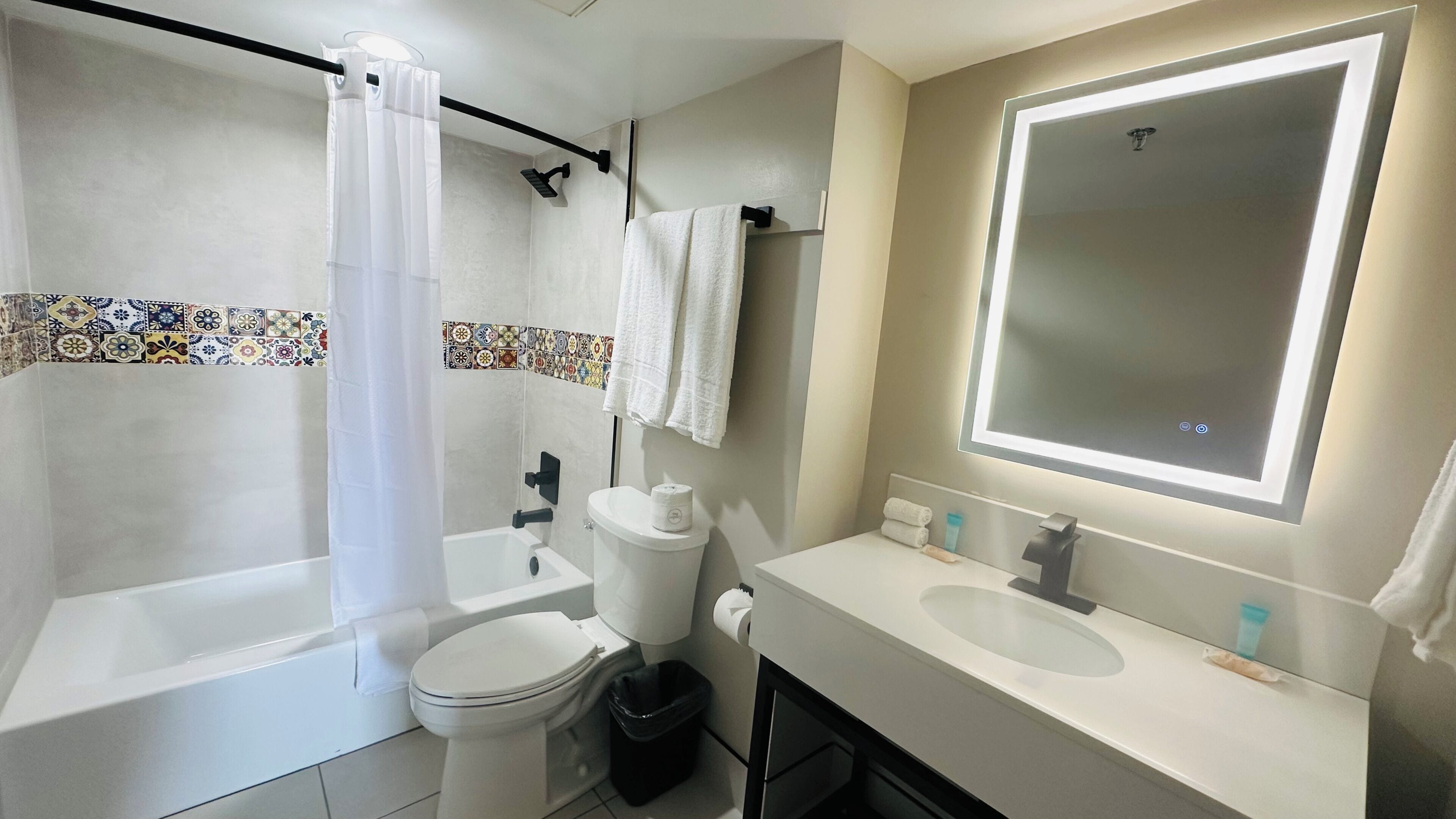 Un baño moderno con bañera, inodoro y lavamanos, que incluye un cortinón de ducha, un perchero para toallas y un espejo grande.