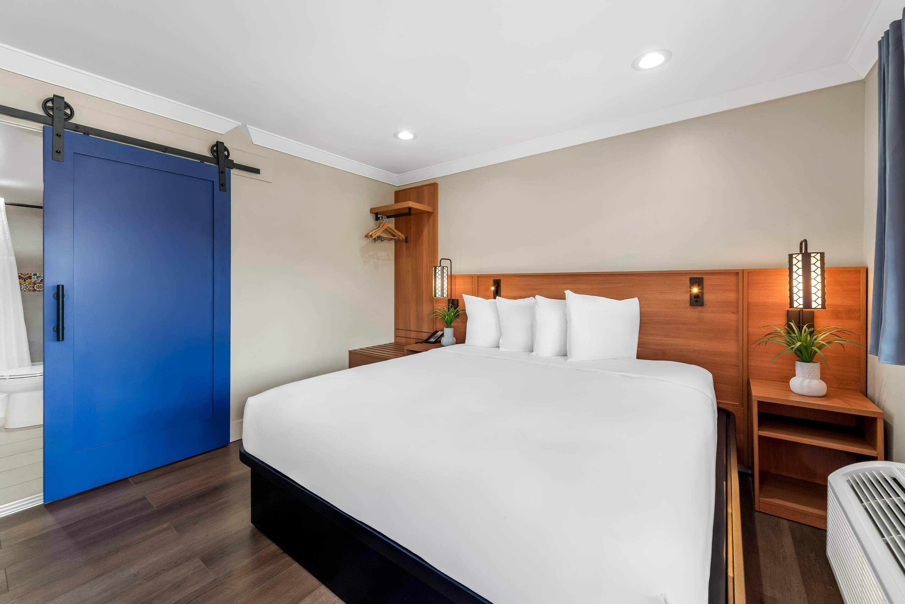 Une chambre d’hôtel avec un grand lit, une tête de lit en bois, des tables de chevet, une porte bleue et un luminaire fixé au mur.