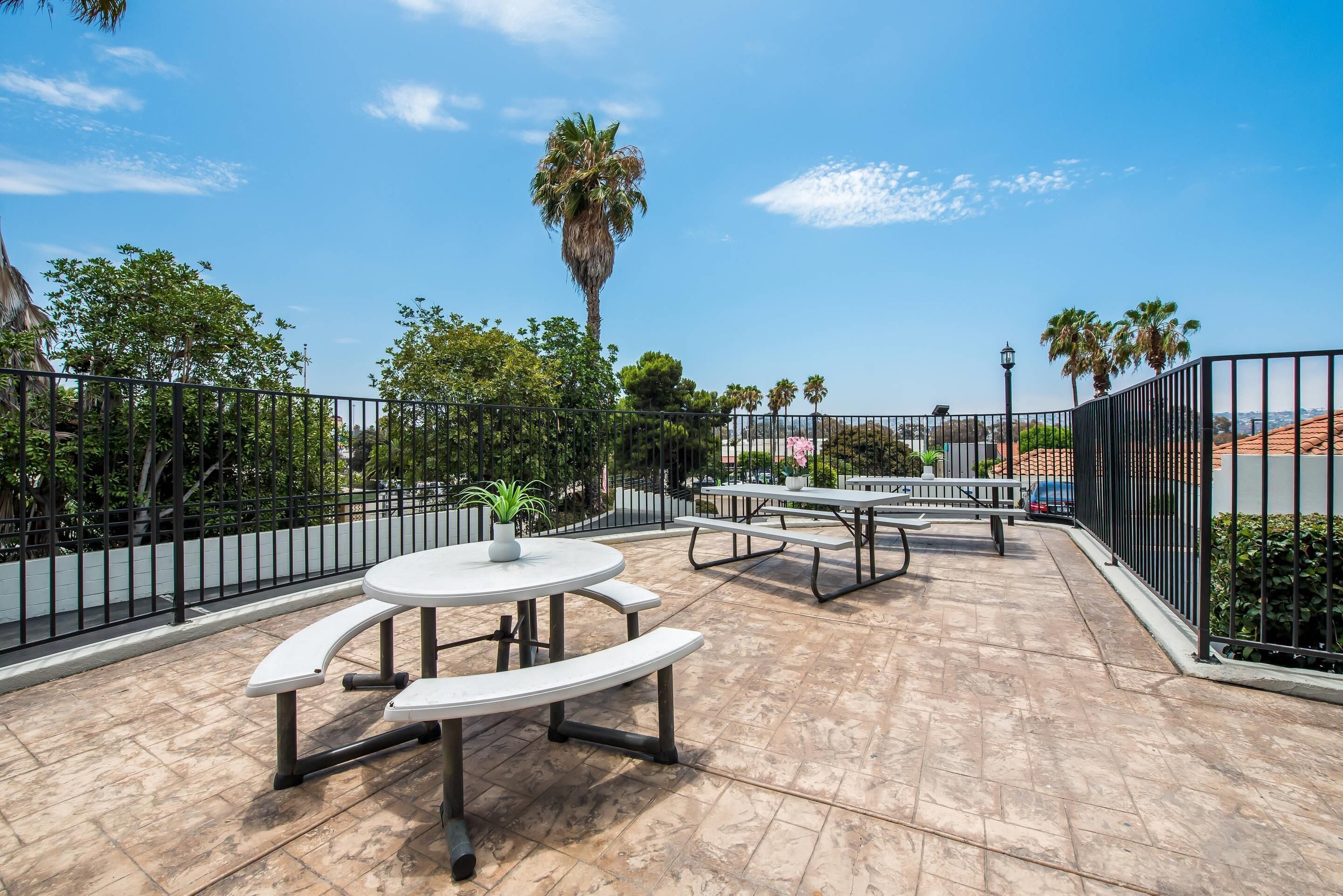 An outdoor patio area with round tables and benches, surrounded by a metal fence, palm trees, and a clear blue sky.