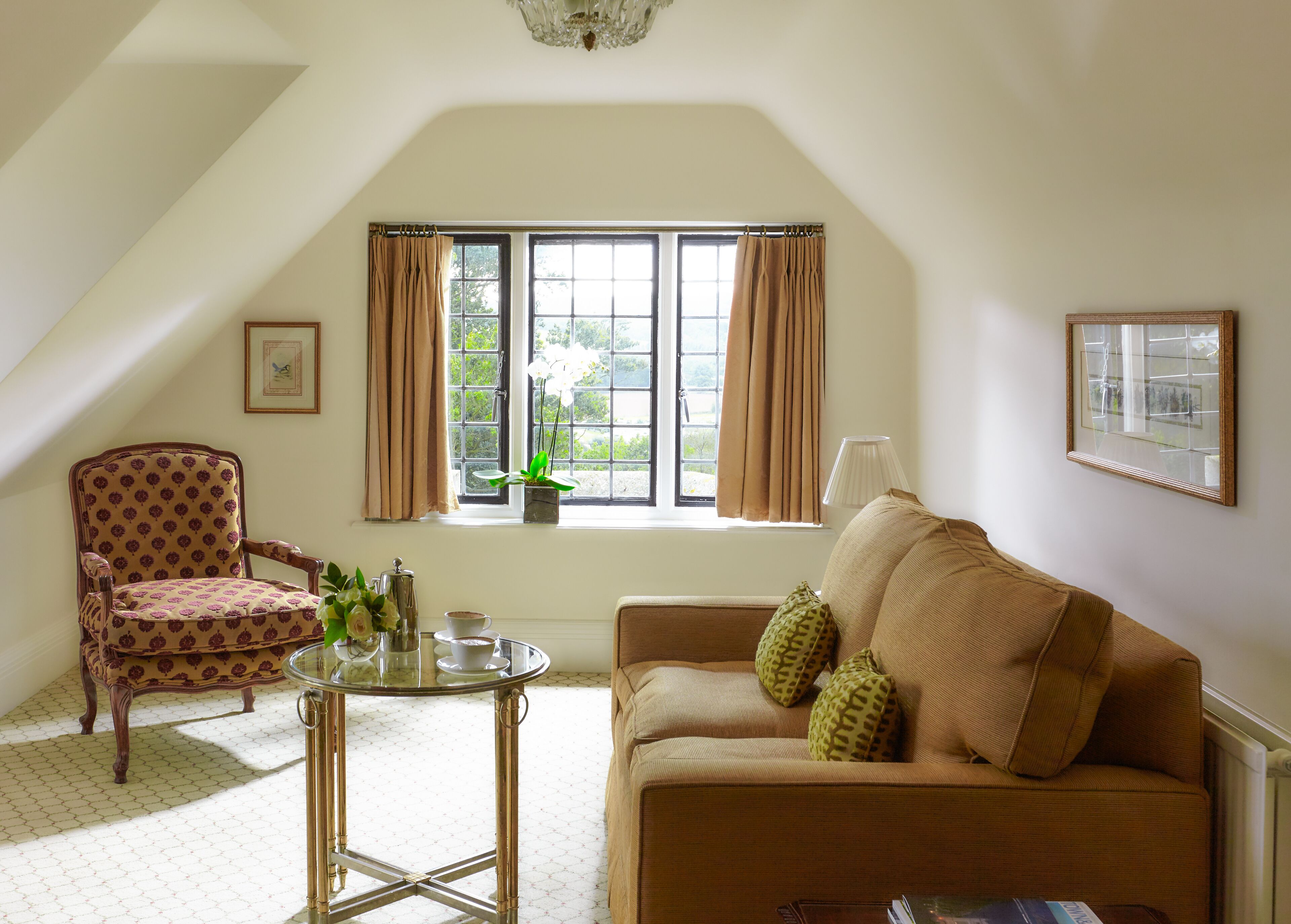 A cozy living room with a high ceiling, a large window with curtains, a patterned armchair, a sofa with decorative pillows, and a small table with a cup and saucer.