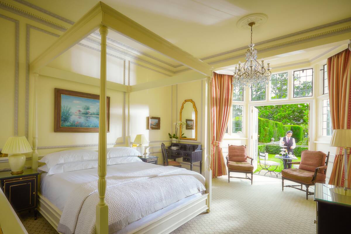 A four-poster bed with a canopy, a desk with a chair, a dining table with chairs, and a view of a garden through large windows.