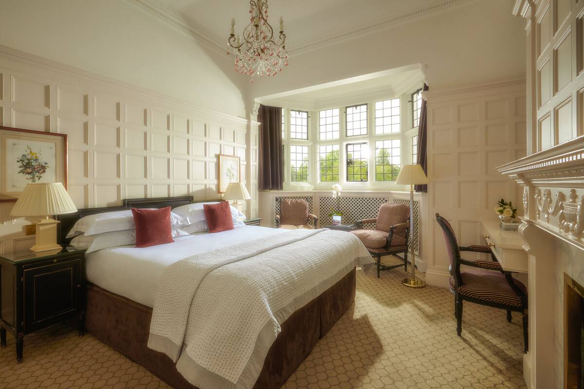 A spacious bedroom with a large bed, a sitting area with chairs, a fireplace, and a view of greenery through the windows.