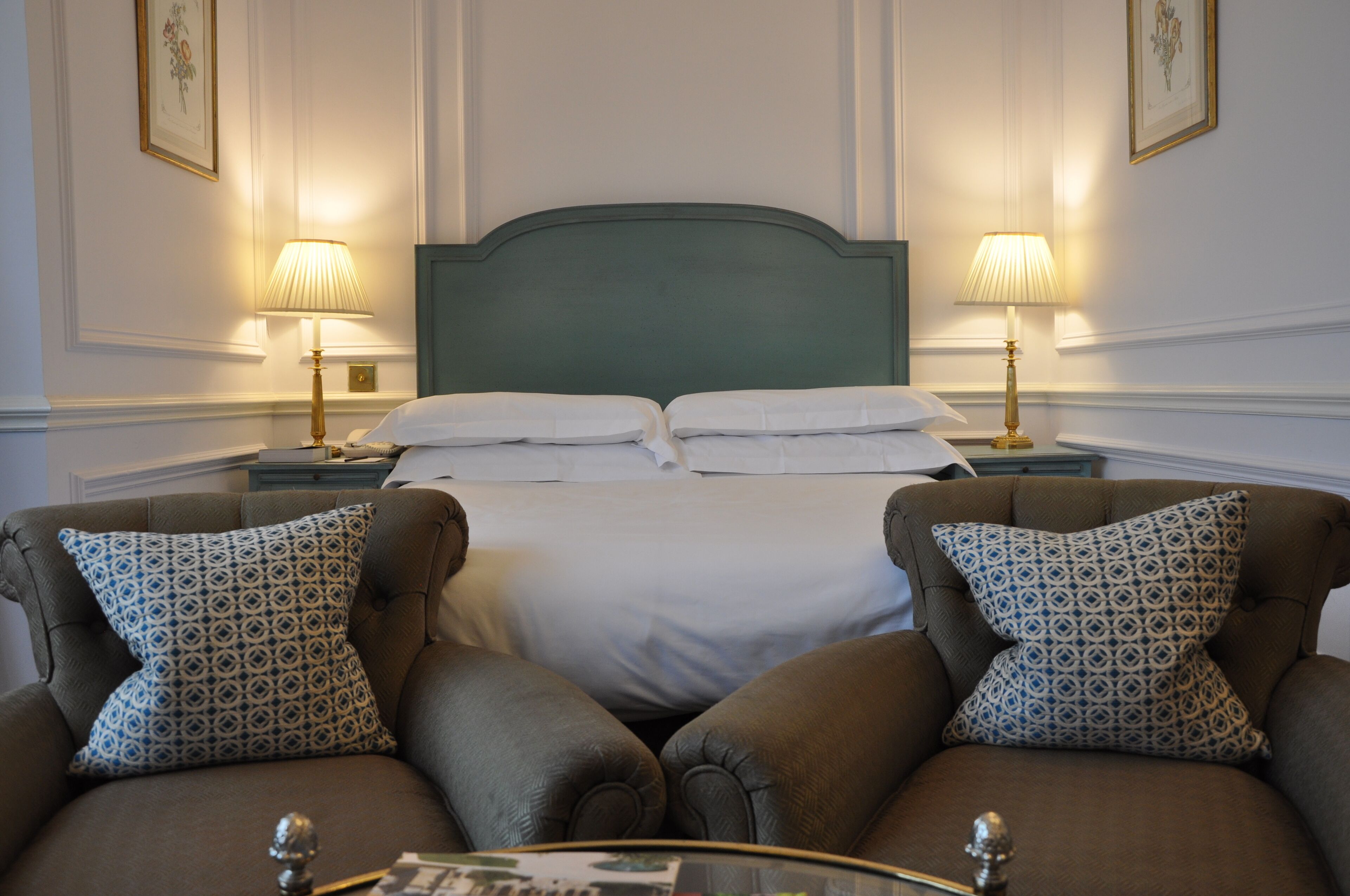 A hotel room with a large bed, two bedside tables with lamps, and two brown armchairs with patterned cushions.
