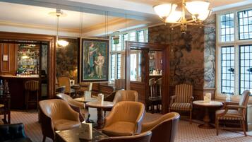 A traditional hotel lobby with leather armchairs, a bar area, and a large painting on the wall.