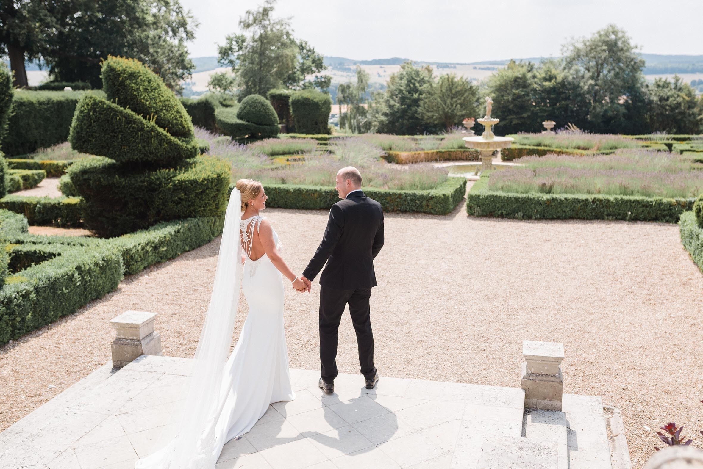 A couple holding hands in a garden with neatly trimmed hedges and a fountain.