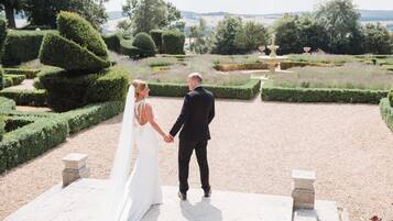 A couple holding hands in a garden with neatly trimmed hedges and a fountain.