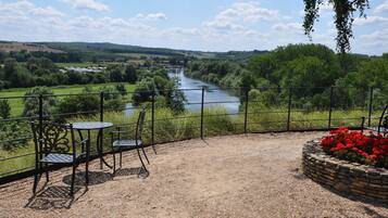 A patio with a view of a river, surrounded by greenery and a stone flower bed.