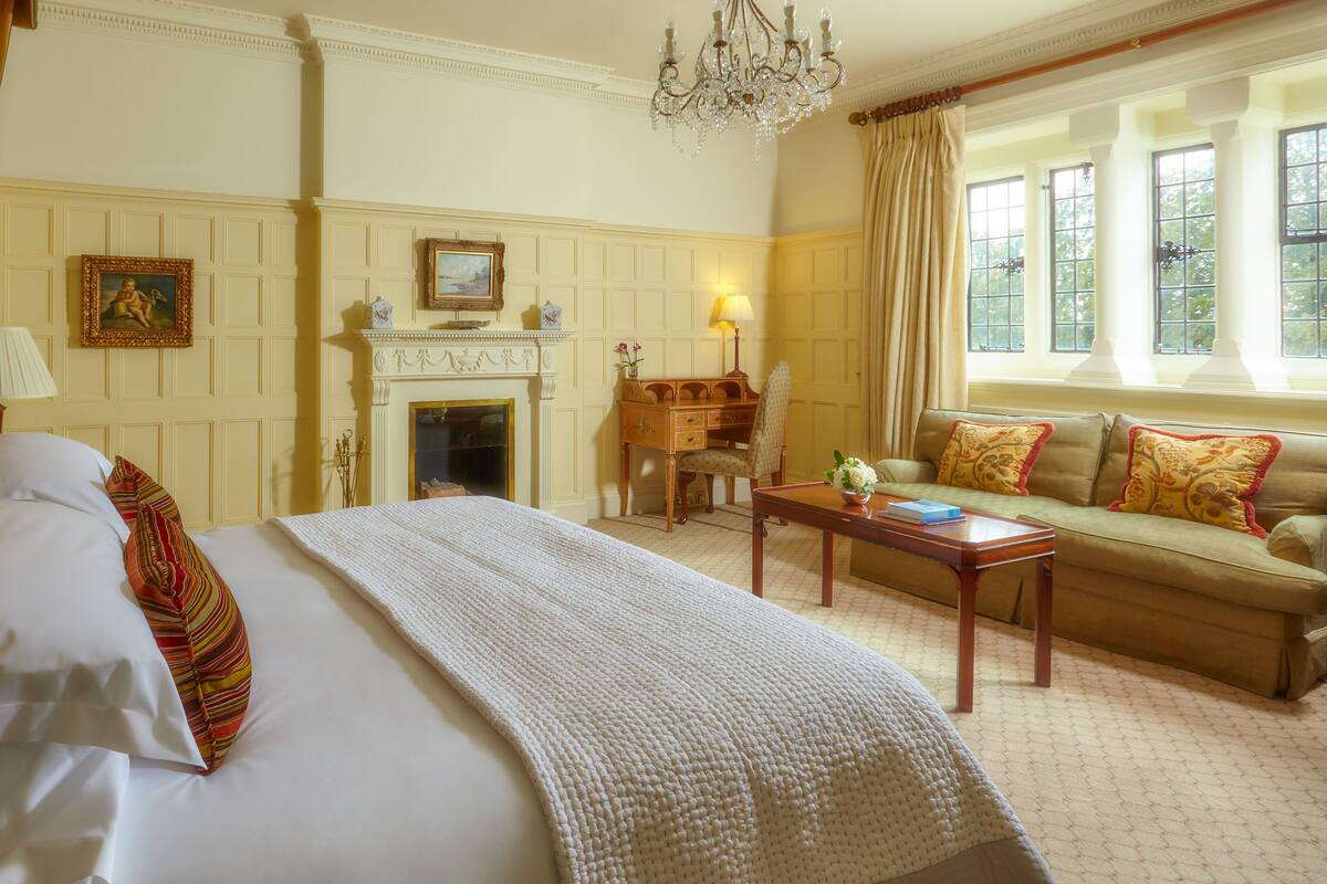 A hotel room with a bed, a sofa, a wooden desk, a fireplace, and large windows.