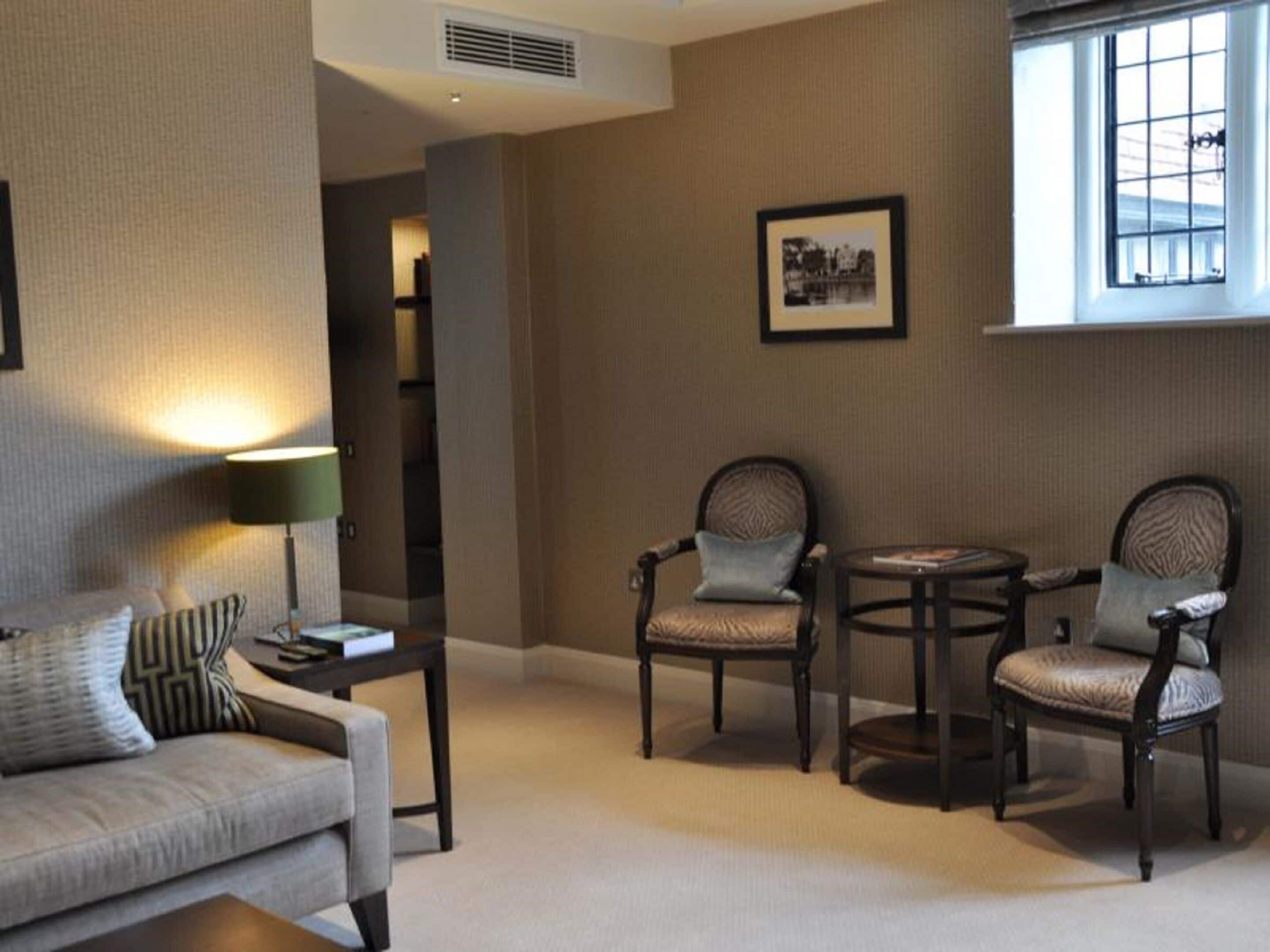 Una sala de estar acogedora con un sofá, dos sillones, una mesita y una lámpara. Hay un cuadro en la pared y una ventana con persianas.