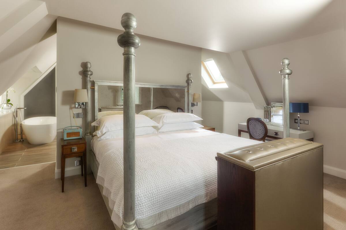 A bedroom with a large bed, a desk, a chair, a vanity with a mirror, and a skylight.