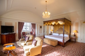 A spacious bedroom with a four-poster bed, a television, a seating area with chairs, and a large window with curtains.